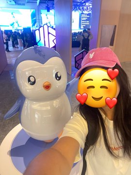 Selfie of a person with a heart-eyes emoji face wearing a pink cap next to a cute penguin mascot statue.