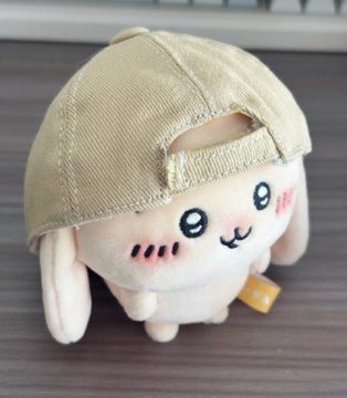 Photograph of a cute plush toy of a white dog-like character wearing a beige baseball cap backward, with a small smile and rosy cheeks.