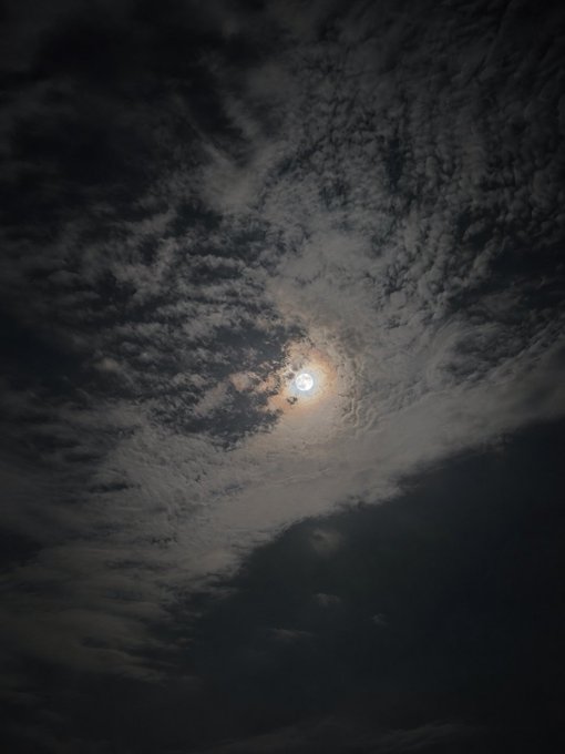 A nighttime photograph showing a bright full moon partially obscured by scattered, dark clouds.