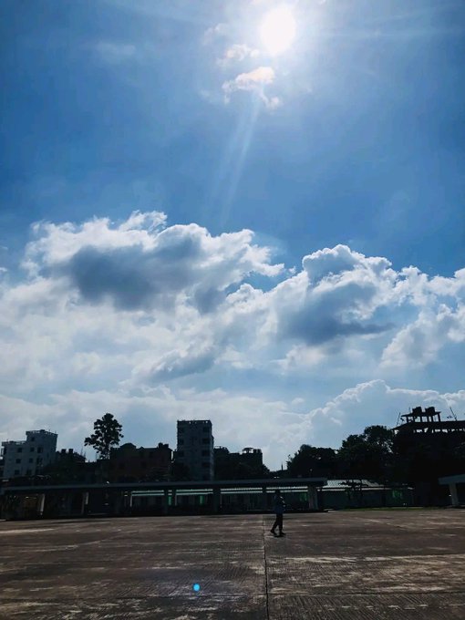 A lone figure stands on a vast empty rooftop beneath a brilliant sun and dramatic cloud-filled sky, embodying the midday energy and forward momentum reflected in the post's "Good Noon" greeting to the #XFamily community. The expansive open space and bright sunlight visually reinforce the message of staying active, growing together, and making the day count.