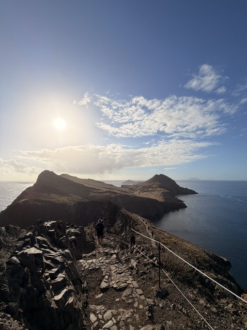 A hiker climbs a steep, rocky coastal trail toward a dramatic volcanic island under a bright morning sky. The “Good morning. Drop your SOL wallet then check DMs.” post contrasts with the serene, scam-resistant reminder implied by the image: pause before taking a risky financial action.