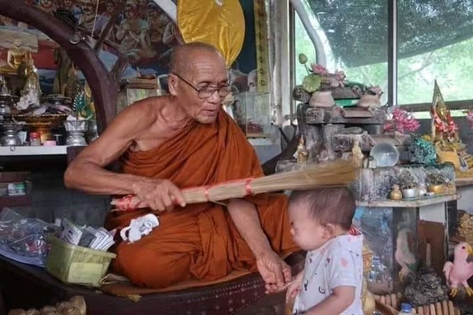 A Buddhist monk in orange robes gently striking a baby with a broom inside a temple filled with religious artifacts and offerings.
