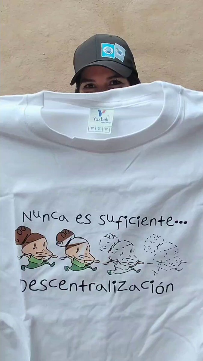 The image features a man wearing a white t-shirt with a drawing of two cartoon characters, one of which has a hat on their head. The man is standing against a beige wall or surface, and there are no other people visible in the picture. The text "NUNCA ES SUFICIENTE... DESCENTRALIZacion" can also be seen in the image.