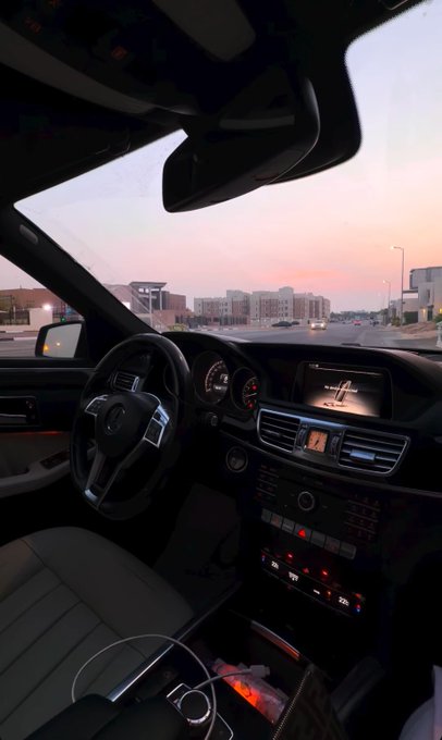 The interior of a car at dusk shows the dashboard, steering wheel, and center console with illuminated controls, looking out through the windshield at a street scene with a sunset sky and distant buildings.