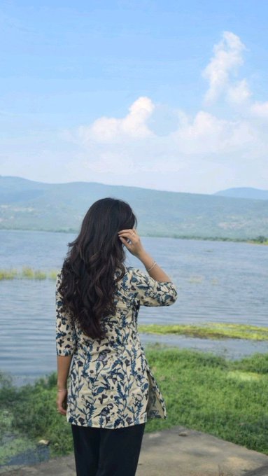 A young woman with long dark hair, wearing a blue and white floral patterned tunic, stands with her back to the camera, looking out over a lake towards distant hills under a cloudy blue sky.