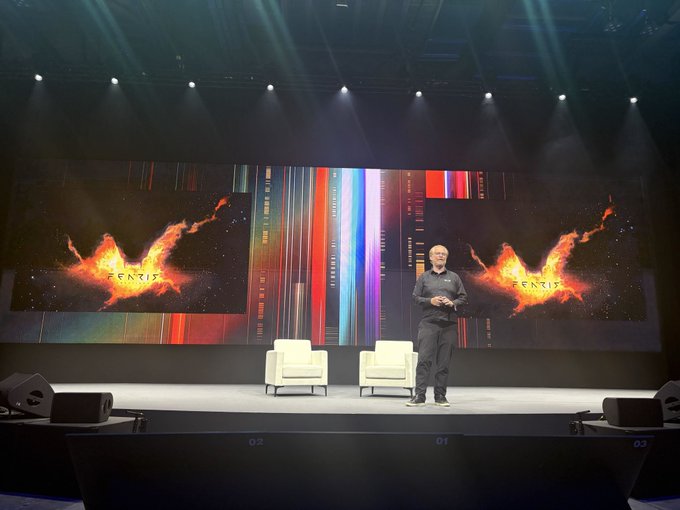 A man stands on a stage during a Fenix presentation, with two empty white chairs behind him and large screens displaying fiery phoenix graphics with 'FENIX' branding.