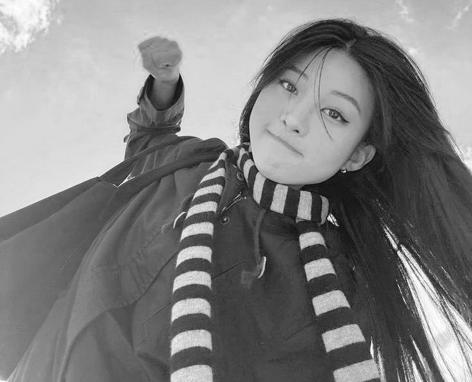 A black and white photo of a young woman with long dark hair wearing a striped scarf and dark jacket, taking a low-angle selfie with her fist raised.