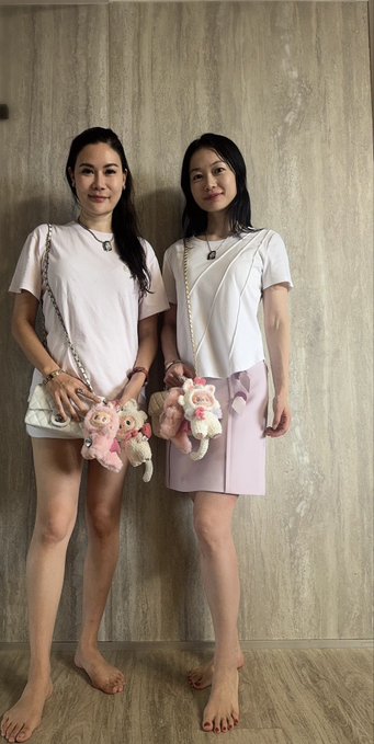 Two women stand side by side against a wooden wall, wearing white t-shirts and carrying matching stuffed animal crossbody bags; one wears white shorts while the other wears a pink skirt.