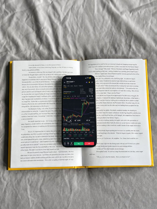 A smartphone showing a BTC/USDT trading chart on a candlestick app rests on the open pages of a paperback novel lying on a white sheet.