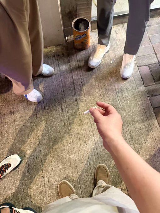A first-person view shows a hand holding a cigarette while standing on a street with several other people's feet and legs visible around them near an Argo tea can.