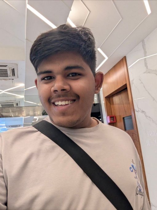 Shriyash, a 20-year-old from India, smiles brightly in what appears to be a hotel or office lobby with marble walls and recessed ceiling lighting, with a crossbody bag strap visible across his beige shirt. The image accompanies his post celebrating one year of growth since attending ETHGlobal Delhi, highlighting milestones like his first international trip, 50+ shipped projects, and multiple wins in the blockchain space.
