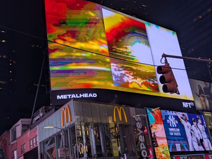 A nighttime photograph of a Times Square billboard displaying a vibrant, glitch-art style NFT titled 'METALHEAD', with a McDonald's restaurant visible below the large screen.