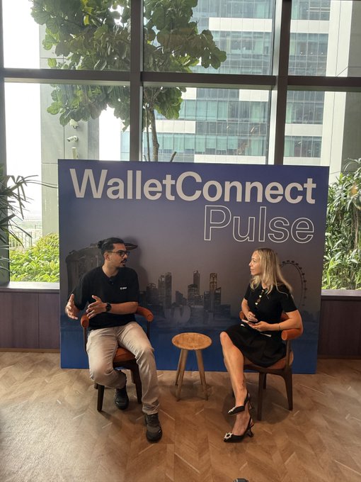 A man and a woman sit on chairs in front of a large blue sign displaying 'WalletConnect Pulse', appearing to engage in a discussion or interview.