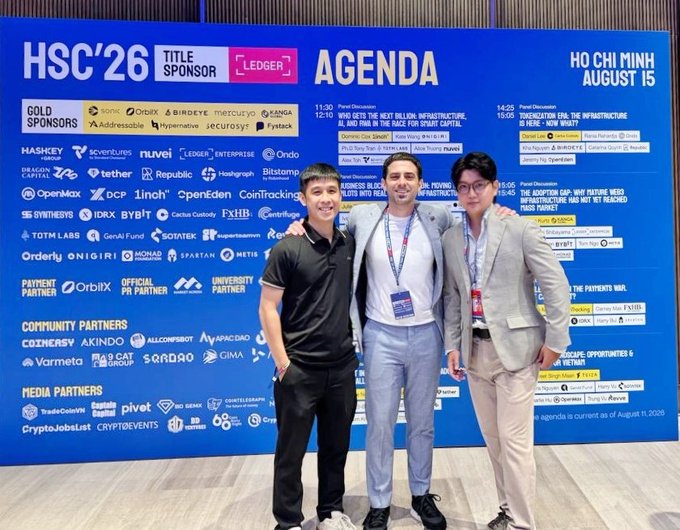 Two men pose together in front of the HSC'26 conference agenda backdrop in Ho Chi Minh on August 15, sponsored by Ledger and featuring brands like Aptos, Circle, and Binance, aligning with the post about attending the Hack Seasons Conference as an Aptos Foundation delegate.
