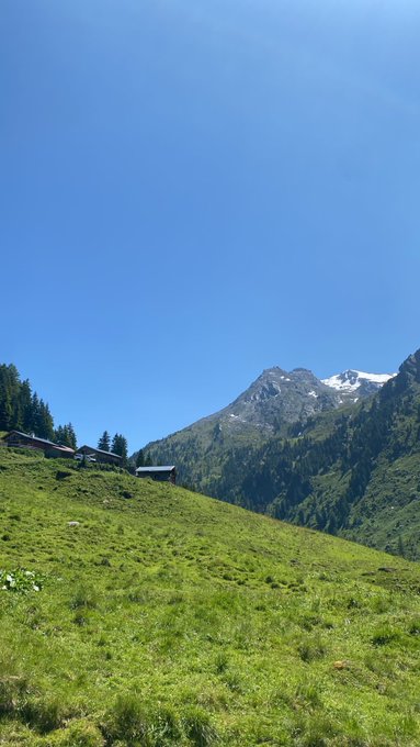 A scenic landscape view of a green mountain meadow with a few small buildings, a forested mountainside, and a snow-capped peak under a clear blue sky.