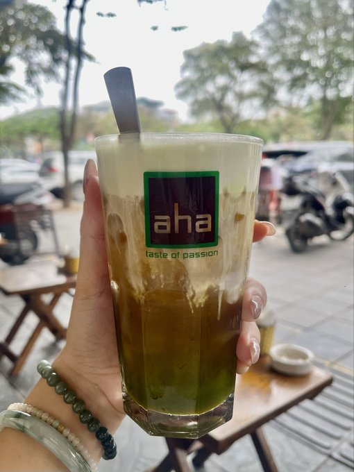The image features a person holding up a tall glass filled with a drink, likely a type of smoothie or milkshake. The drink has a green color that stands out against the brown background. In the background, there are several cars parked outside, indicating that this scene takes place in an urban setting. The post accompanying the image is titled "Xin chào mọi người Em là Thái Thanh đây Thứ có thúc một tuần mới bình an, vui vẻ, nhiều năng lượng tích cực và thật nhiều thành công kết nối". The post seems to be promoting a smoothie or milkshake, possibly from a nearby cafe.