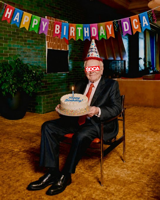 Man in a pinstripe suit and red tie sitting on a chair holding a birthday cake, with a birthday hat and '$DCA' glasses over his eyes, beneath a 'HAPPY BIRTHDAY DCA' banner.