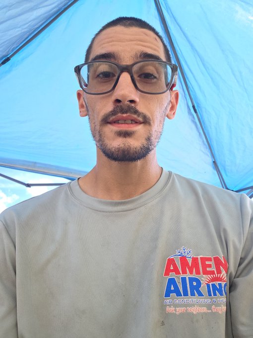 A selfie of a man with short dark hair, glasses, and a goatee wearing a beige sweatshirt with 'AMEN AIR INC' branding. He stands under a blue tarp or tent structure.