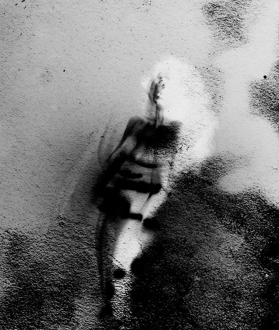 A grainy, black-and-white image shows a blurred silhouette of a woman walking on a wet surface, surrounded by atmospheric mist and dark, textured shadows. The artwork's haunting, indistinct quality visually reinforces the caption's theme of "a woman who doesn't exist," reflecting an AI-generated or digitally crafted character that feels both present and ghostly.