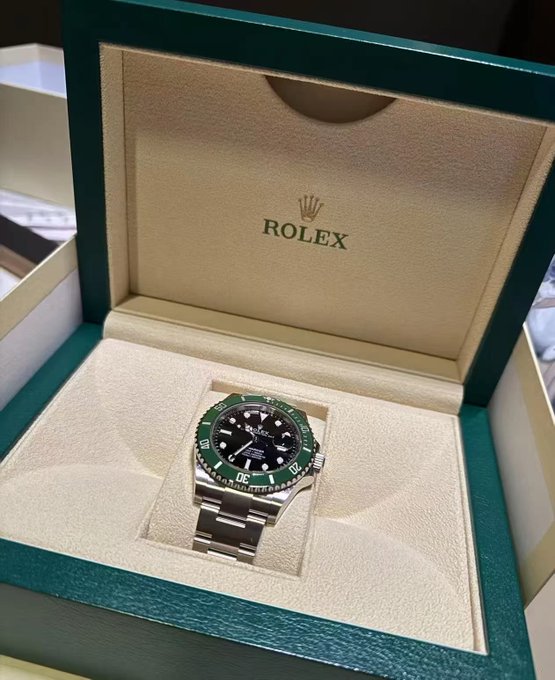 A Rolex Submariner watch with a green bezel and black dial is displayed in its green presentation box.