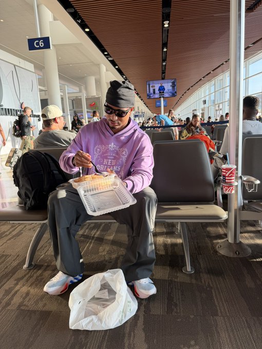 A man in a New Orleans hoodie eats takeout food at gate C6 in an airport terminal, reflecting a casual "Good Morning" travel moment shared by $ANSEM Bulls.
