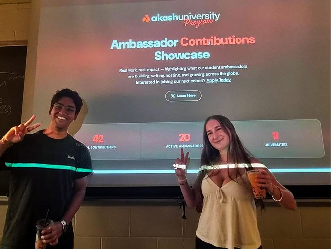 Two people smile and pose with peace signs in front of a projection screen displaying 'Akash University Program - Ambassador Contributions Showcase', which lists 42 contributions, 20 active ambassadors, and 11 universities.