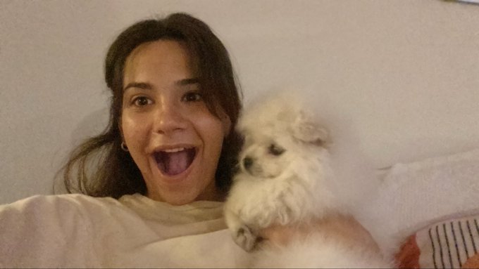 A young woman with long dark hair beams with an excited, open-mouthed expression while cuddling a fluffy white puppy, perfectly capturing the joyful energy and carefree spirit of being 21.