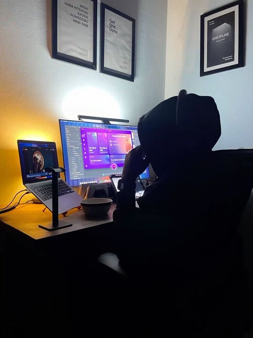 A silhouetted figure in a hoodie sits at a desk with two monitors displaying design software, with framed posters on the wall reading 'NIEMAND HAT DIE ABSICHT EINE MAUER ZU ERRICHTEN POMPADOUR WAGI-SAAR' and 'Get Shit Done' alongside a 'DISCIPLINE' poster.