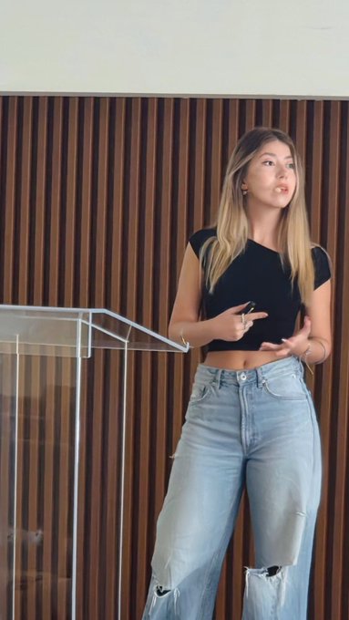 A woman in a black crop top and ripped jeans stands at a clear podium speaking against a wood-slatted wall.