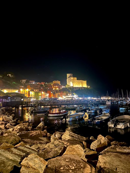 A nighttime view of a coastal Italian harbor features small boats moored in the water, with a brightly lit medieval fortress perched on a hilltop above colorful buildings and restaurants.