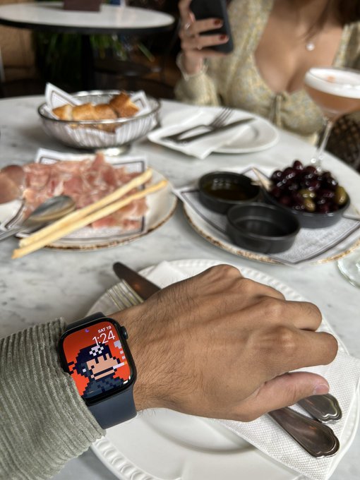 A close-up of a person's hand wearing an Apple Watch with a pixel art design showing 'SAT 124', resting on a plate at a restaurant table set with food and drinks.