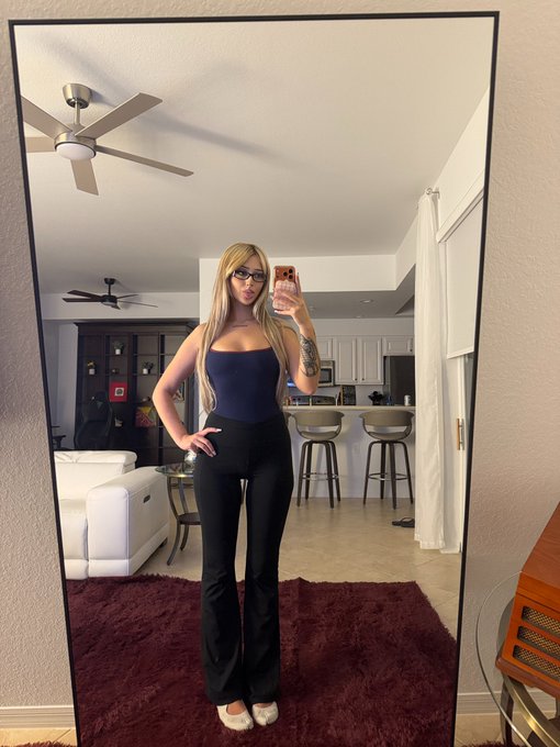 A mirror selfie of a woman with blonde hair, glasses, a blue top, and black pants, standing in a living room with a ceiling fan visible.