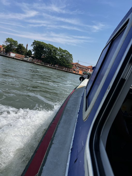 A POV photograph from a blue boat speeding through water in what appears to be Venice, with historic buildings and a brick tower visible along the tree-lined shore.