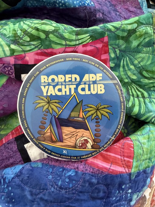 A circular sticker or button labeled 'BORED APE YACHT CLUB' size XL, featuring a stylized beach scene with a yacht and palm trees, rests on a colorful patchwork quilt.