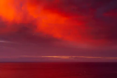 A dramatic sunset or sunrise displays vibrant red and orange clouds over a calm ocean horizon.