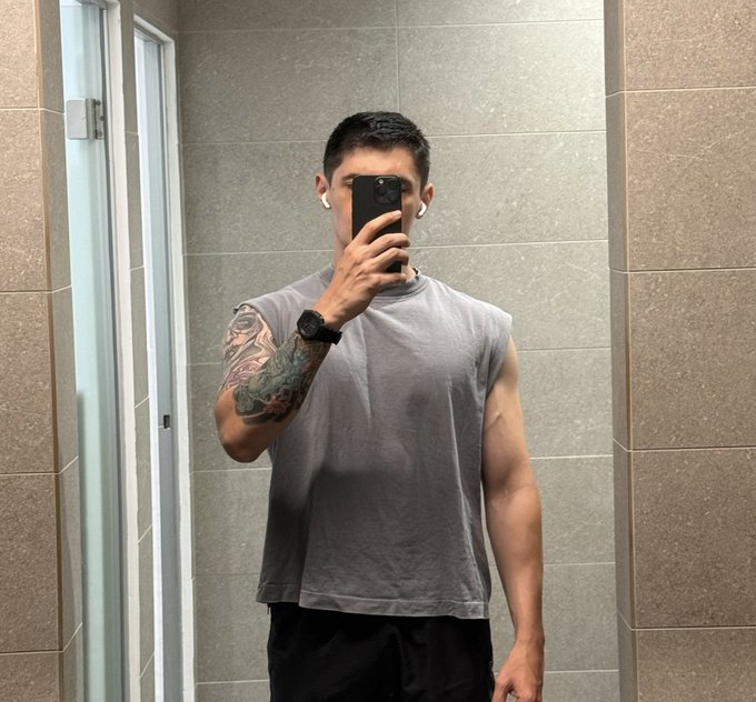 Mirror selfie of a young man in a sleeveless gray shirt with a tattooed arm, wearing a smartwatch and earbuds, standing in a tiled bathroom.