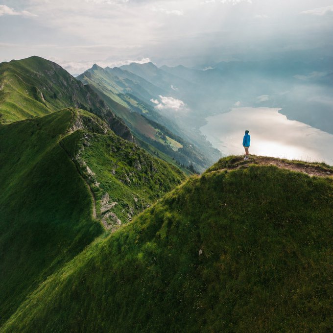 A breathtaking landscape photograph of a person standing on the edge of a lush green mountain ridge, gazing out over a misty valley and a large lake reflecting the sunlight.