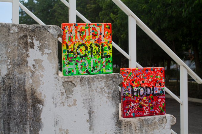 Two small vibrant paintings are displayed on weathered concrete steps; the left one reads "HODL" in green and yellow, while the right one is red, both covered in repeated "HODL" text.