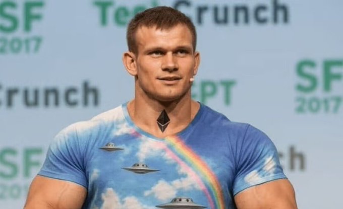 A muscular man with a neck tattoo wearing a sky-themed t-shirt featuring UFOs and a rainbow, standing on a stage with "TechCrunch SF 2017" visible in the background.