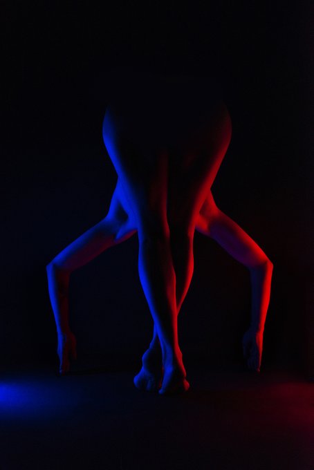 Artistic photo of a person's legs and torso posed dramatically against a dark background, illuminated by contrasting blue and red lighting.