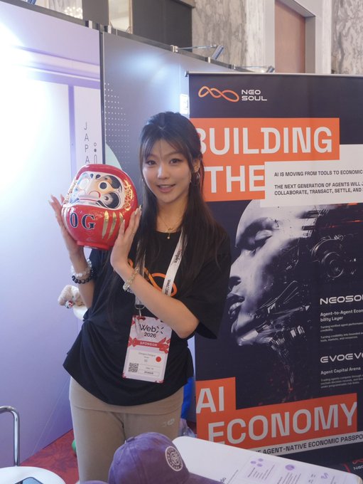 A young woman at the WebX 2020 conference in Japan holds a red Daruma doll, standing beside a Neo Soul booth banner promoting 'BUILDING THE AI ECONOMY' and Agent-to-Agent Ecosystems.