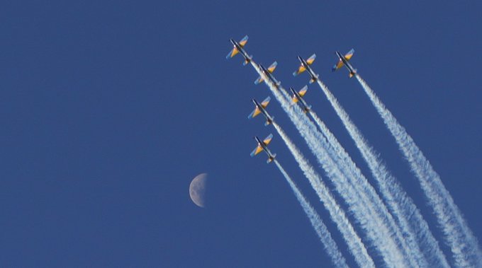 A formation of jets flying in the sky leaving white smoke trails, with a crescent moon visible in the deep blue background.