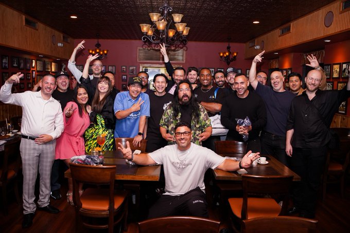 A large group of people poses enthusiastically together at a wooden table in a warmly lit restaurant or bar with a chandelier hanging above.