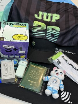 The image features a black table with various items scattered across its surface. On the table, there are two stuffed animals - one white cat toy and another blue dog toy. A passport holder can also be seen among the items. The main focus of the scene is a limited edition CatLumpur merchandise item that includes a passport holder and a mini handheld fan. The text "JUP 26" is visible on the merchandise, indicating its association with the popular Japanese soccer team, Jun-use.