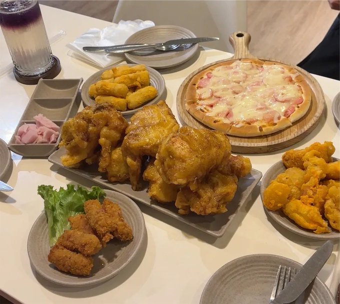 A spread of fried foods including a bacon-topped pizza, crispy fried chicken, onion rings, sweet potato fries, and fried chicken cutlets, paired with a purple layered drink. The image illustrates a hearty meal meant to fuel a full afternoon of work ("cào X" — scrolling X) after lunch, reflecting the post's casual tone about indulging in food before diving back into social media.