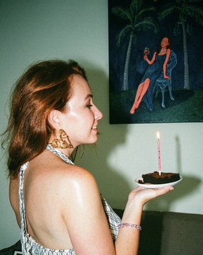 A woman in a sleeveless top holds a small chocolate cake with a single lit candle, standing near a painting of a seated figure.