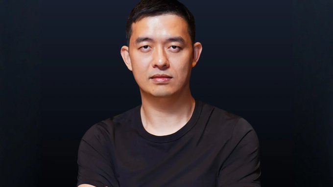 A professional headshot of an East Asian man in a black t-shirt against a dark background.