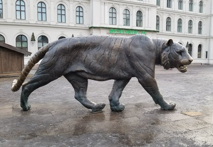 A large bronze tiger statue stands in a courtyard in front of a white building with arched windows, with green text reading 'FINNAUST' partially visible in the background.