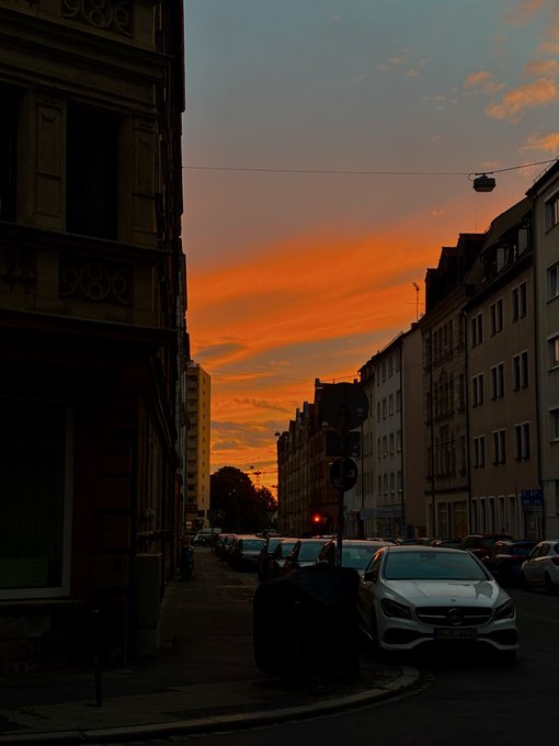 A street view at sunset, where the sky glows with vibrant orange and blue hues above darkened silhouettes of buildings, parked cars, and a streetlamp on a city block.