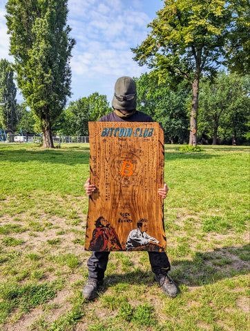 The image features a man standing in a grassy field, holding up an old wooden sign with graffiti on it. The sign has the words "Butterfly Club" written on it. The background of the photo shows trees and a park setting, indicating that this scene takes place outdoors.